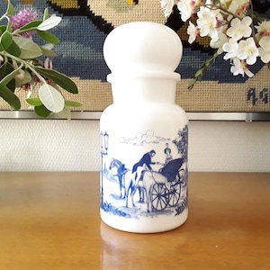 Peut inclure: Un pot à pharmacie blanc avec un dessus arrondi et un motif bleu et blanc représentant une calèche. Le pot est exposé sur une surface en bois, avec des fleurs en arrière-plan.