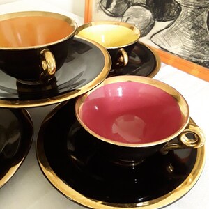 May include: A set of black teacups and saucers with gold trim. The interior of the cups are in shades of pink, yellow, and orange. The teacups are on a white surface. A framed black and white drawing is in the background.