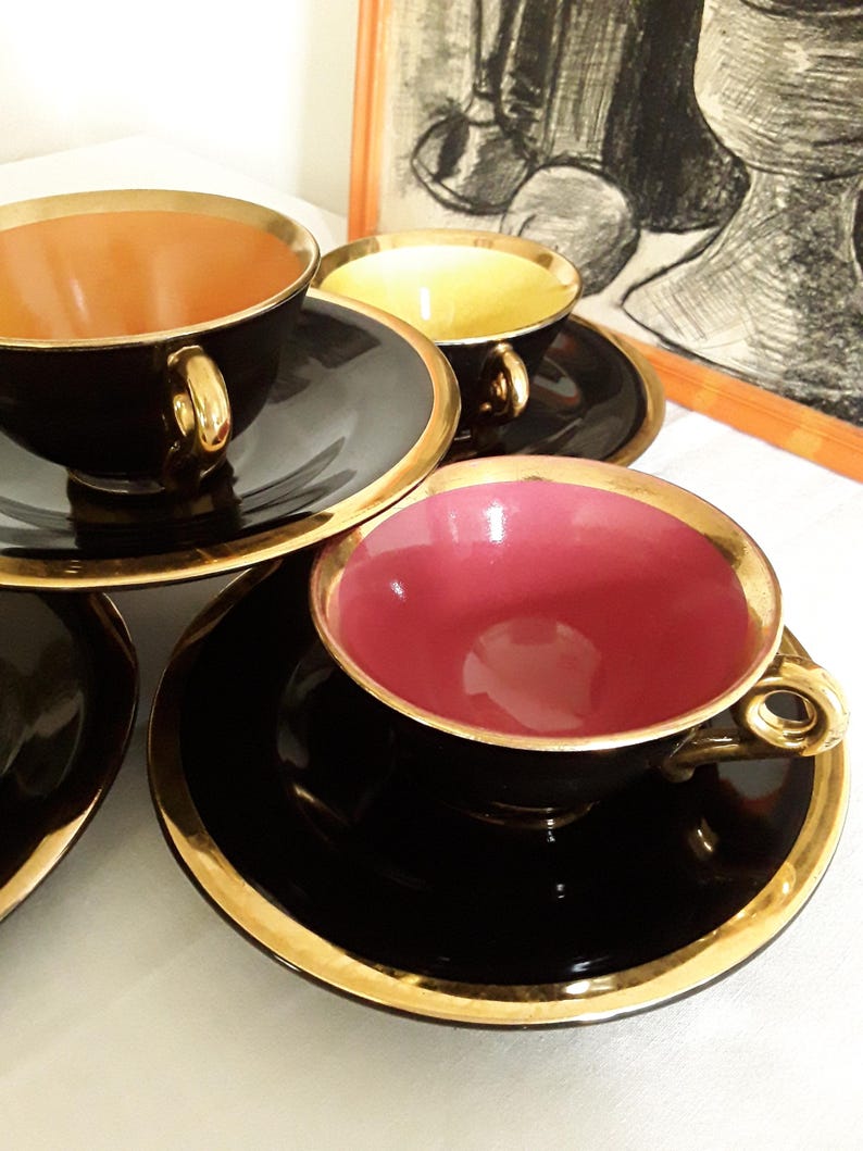 May include: A set of black teacups and saucers with gold trim. The interior of the cups are in shades of pink, yellow, and orange. The teacups are on a white surface. A framed black and white drawing is in the background.