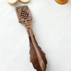 Puede incluir: Un cascanueces de madera con un diseño de uva tallado. El cascanueces está cerrado y tiene un acabado marrón.