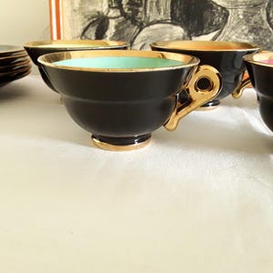 May include: A set of black teacups with gold trim and handles. The interior of the cups features different colors, including teal, orange, and pink. A stack of black saucers with gold trim is visible in the background. The teacups are arranged on a white surface.