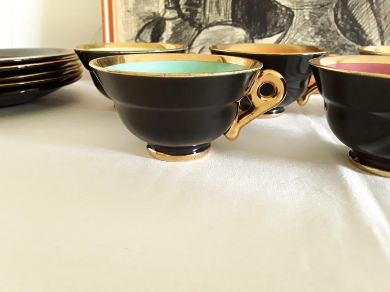 May include: A set of black teacups with gold trim and handles. The interior of the cups features different colors, including teal, orange, and pink. A stack of black saucers with gold trim is visible in the background. The teacups are arranged on a white surface.