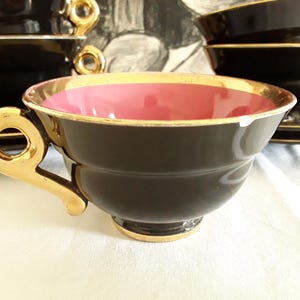May include: A set of black teacups with gold trim and handles. The interior of the cups is a vibrant pink. Saucers are visible in the background. The teacups are arranged on a white surface.