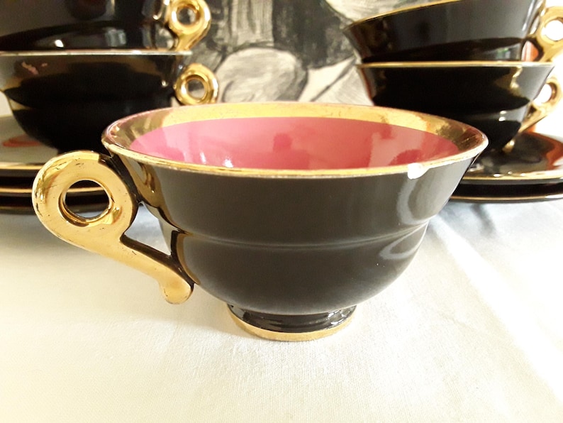 May include: A set of black teacups with gold trim and handles. The interior of the cups is a vibrant pink. Saucers are visible in the background. The teacups are arranged on a white surface.