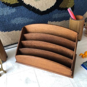 Puede incluir: Un organizador de escritorio de madera con cinco ranuras, ideal para guardar papeles o correo. El organizador está hecho de madera de tonos cálidos y se encuentra sobre una superficie blanca. También se ve una pequeña figura de gato de latón y una pequeña figura de gato naranja.