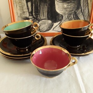 May include: Set of vintage black teacups and saucers with gold trim. The teacups have colored interiors in shades of pink, green, and orange. The saucers and cups are arranged on a white surface, with a decorative bottle and framed artwork in the background.