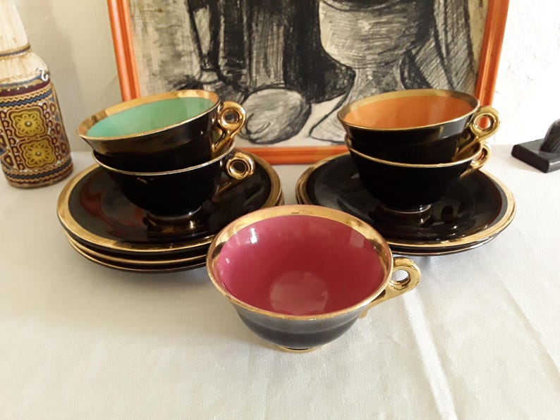 May include: Set of vintage black teacups and saucers with gold trim. The teacups have colored interiors in shades of pink, green, and orange. The saucers and cups are arranged on a white surface, with a decorative bottle and framed artwork in the background.