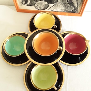 May include: A set of five colorful teacups and saucers. Each cup is a different color: yellow, orange, pink, green, and light green. The cups have gold handles and sit on black saucers with gold rims. An artwork is in the background.