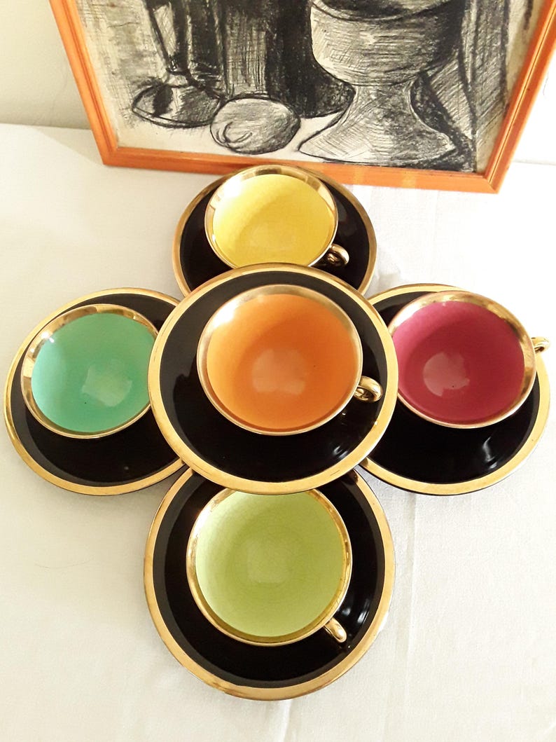 May include: A set of five colorful teacups and saucers. Each cup is a different color: yellow, orange, pink, green, and light green. The cups have gold handles and sit on black saucers with gold rims. An artwork is in the background.