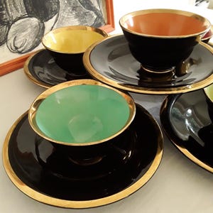May include: A collection of vintage teacups and saucers. The cups have black exteriors with gold rims and colorful interiors in yellow, orange, and green. The saucers are black with gold trim. An artwork is in the background.