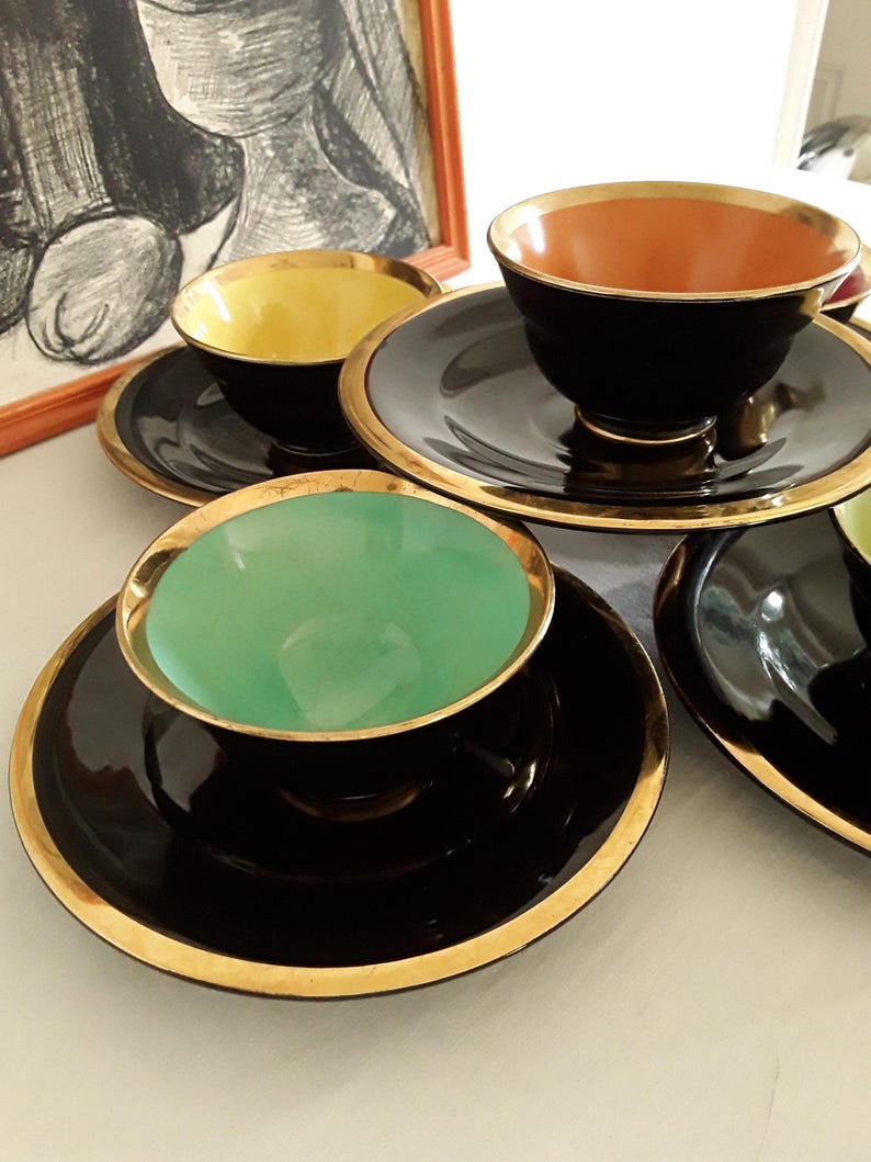 May include: A collection of vintage teacups and saucers. The cups have black exteriors with gold rims and colorful interiors in yellow, orange, and green. The saucers are black with gold trim. An artwork is in the background.