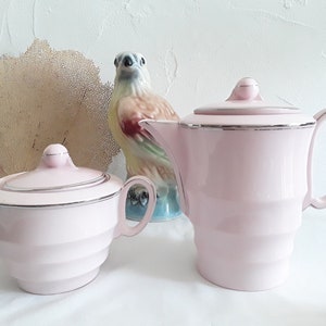 Puede incluir: Una tetera y un azucarero de cerámica rosa con ribetes plateados. La tetera tiene un cuerpo redondeado y una tapa con un pomo. El azucarero tiene una tapa con un pomo y dos asas.