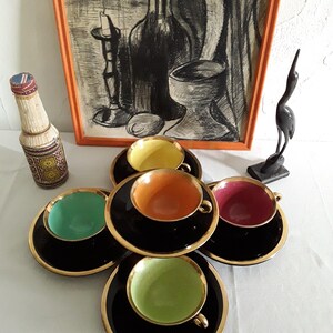 May include: A set of six vintage teacups and saucers. The cups have black exteriors with gold rims and colorful interiors in shades of yellow, orange, green, and red. The saucers are black with gold trim. A framed artwork and decorative items are in the background.