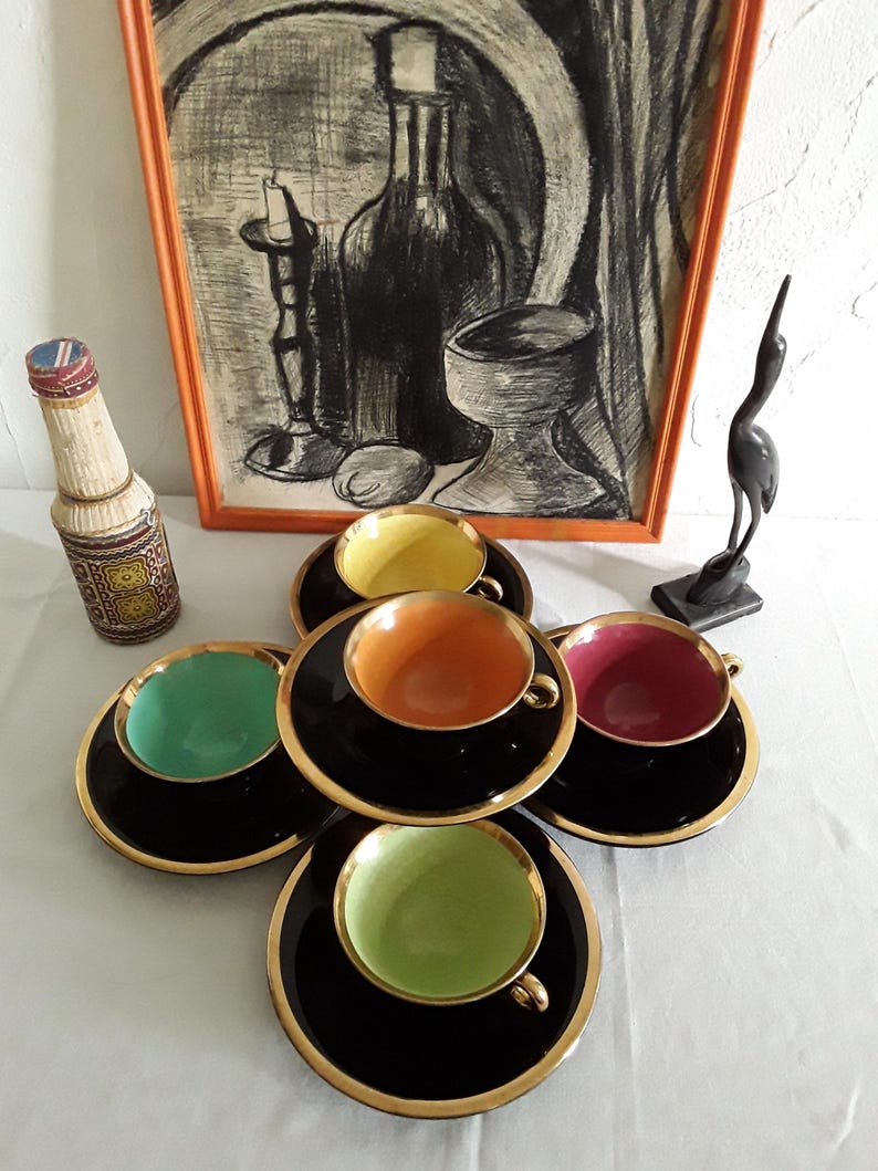 May include: A set of six vintage teacups and saucers. The cups have black exteriors with gold rims and colorful interiors in shades of yellow, orange, green, and red. The saucers are black with gold trim. A framed artwork and decorative items are in the background.