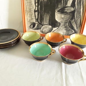 May include: A set of vintage teacups and saucers. The cups are black with gold trim and feature interiors in various colors: green, orange, yellow, teal, and pink. A stack of black saucers with gold rims is also present.