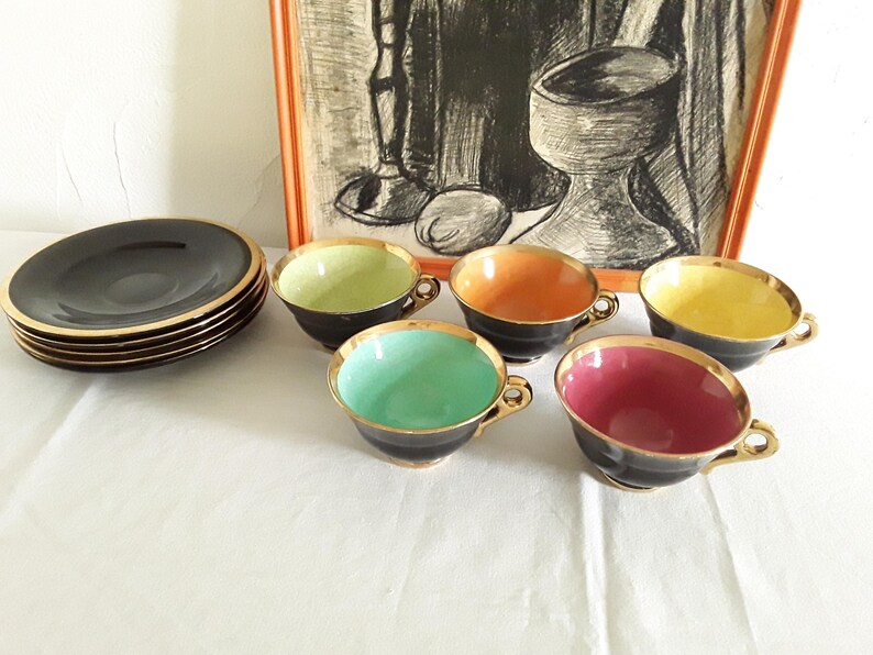 May include: A set of vintage teacups and saucers. The cups are black with gold trim and feature interiors in various colors: green, orange, yellow, teal, and pink. A stack of black saucers with gold rims is also present.