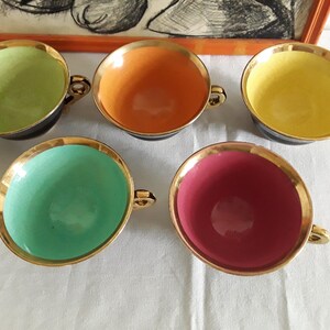 May include: Set of five colorful teacups with gold rims and handles. The interior colors include green, orange, yellow, teal, and pink. The cups have a black exterior and sit on a white surface. A framed artwork is in the background.