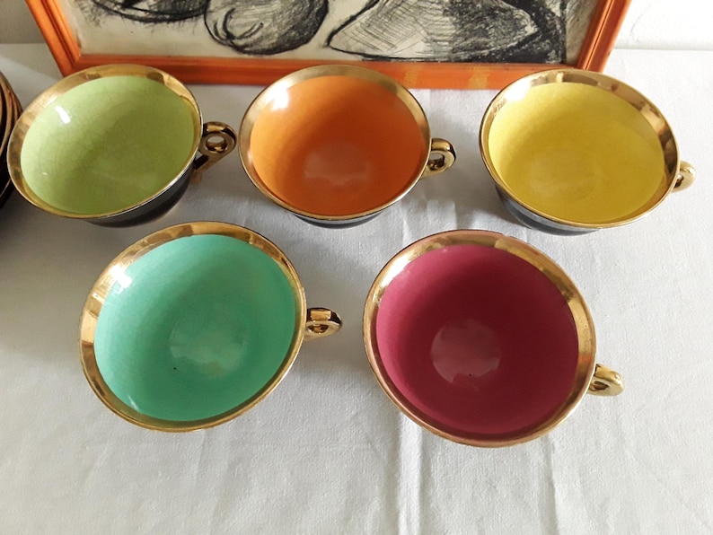May include: Set of five colorful teacups with gold rims and handles. The interior colors include green, orange, yellow, teal, and pink. The cups have a black exterior and sit on a white surface. A framed artwork is in the background.