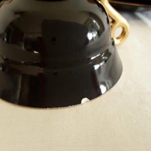 May include: Close-up of a black teacup with a gold handle. The cup has a glossy finish and a small chip on the rim. The handle is a curved design. The teacup is part of a tea set.