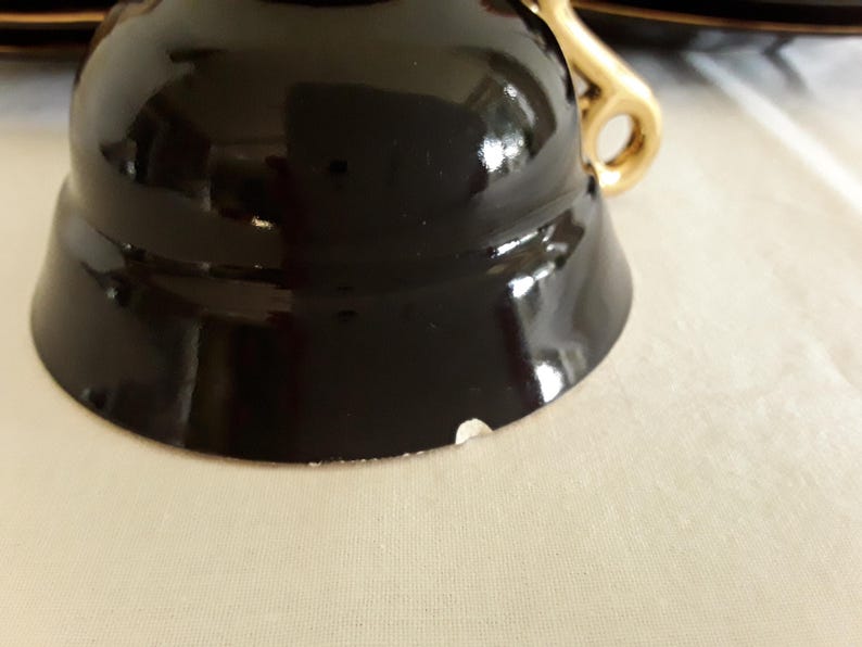 May include: Close-up of a black teacup with a gold handle. The cup has a glossy finish and a small chip on the rim. The handle is a curved design. The teacup is part of a tea set.