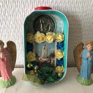 May include: A religious shrine made from a repurposed tin. The interior features a religious image, a metal medallion, and is decorated with yellow and blue flowers, green beads, and a green ribbon. Two small angel figurines flank the shrine.