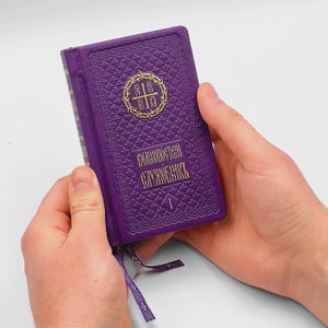 Puede incluir: Un libro de tapa dura morado con superficie texturizada y letras doradas. La portada presenta un emblema circular con una cruz dentro de una corona de espinas, el texto cirílico "ВЕЛИКОПОСТНЫЙ СЛУЖЕБНИКЪ" y el número "1". Un marcapáginas de cinta morada cuelga de la parte inferior.