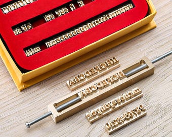 May include: A set of brass letter stamps and a stamp holder on a wooden surface. The stamps are arranged in a red-lined box and are also placed in the stamp holder. The letters are in various sizes and fonts.