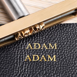 Könnte beinhalten: Ein Messing-Brennstempel mit Gewindestange und verstellbaren Buchstaben, die ADAM bilden, platziert auf einer schwarzen, strukturierten Lederoberfläche, auf der der Name ADAM zweimal in Gold in einer gestapelten Anordnung eingeprägt ist.