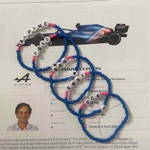 Puede incluir: Un brazalete de cuentas azul y rosa con las palabras "Alpine", "Gas", "Team", "Stats", "E03", "OCO" y "PGIO" escritas en él. El brazalete está sobre un fondo blanco con un patrón de cuadrícula. El texto "Alpine F1® Team" está en la parte superior de la imagen.