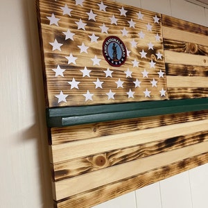 May include: A wooden American flag with a burnt finish. The flag is mounted on a wall and has a green shelf above it. The flag has a white star design and a red, white, and blue Army National Guard emblem in the center.