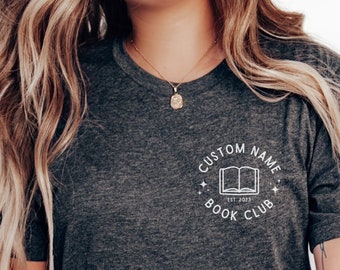 May include: A dark grey t-shirt with a white graphic of a book and the text "Custom Name Book Club Est. 2023".