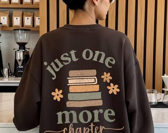 May include: A dark brown sweatshirt featuring the words "just one more chapter" and a graphic of stacked books. The design includes flower motifs. The sweatshirt is worn by a person with a white hair clip and sunglasses.