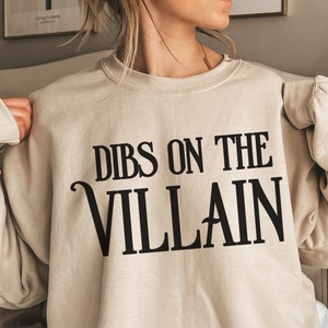 K&ouml;nnte beinhalten: Beiges Sweatshirt mit dem schwarzen Text "DIBS ON THE VILLAIN", eine verspielte und k&uuml;hne Aussage. Das Sweatshirt hat eine neutrale Farbe und eignet sich f&uuml;r Freizeitkleidung. Der Text ist in einer stilisierten Schriftart gehalten und verleiht dem Kleidungsst&uuml;ck eine pers&ouml;nliche Note.