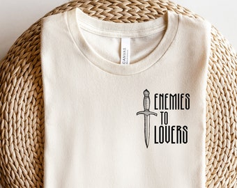 May include: Cream-coloured t-shirt featuring the text "ENEMIES TO LOVERS" and a dagger graphic. The shirt is made of a soft material and is displayed on a woven, circular surface. The text and graphic are in black.