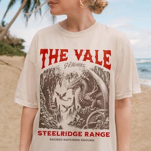 Peut inclure: Une femme portant un t-shirt blanc avec un imprimé graphique d'un dragon et le texte "The Vale Dragons Steelridge Range Sacred Hatching Ground".