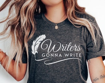 May include: A dark heather gray short-sleeve t-shirt featuring a white graphic of a quill pen and the text "Writers GONNA WRITE" printed across the chest. The shirt has a crew neckline and is paired with light blue denim jeans.