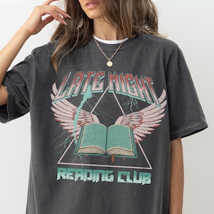 May include: Dark gray t-shirt with a graphic design. The design features the text "LATE NIGHT READING CLUB" with a book, wings, and lightning bolts. The shirt has short sleeves and a relaxed fit.