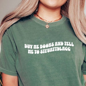 May include: Light green t-shirt with the text "BUY ME BOOKS AND TELL ME TO STFUATTDLAGG" in white, a round gold necklace, and a gold bracelet. The shirt is a solid color and has short sleeves.