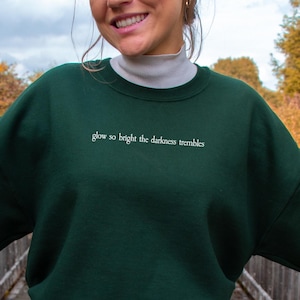 May include: A dark green sweatshirt with the white text "glow so bright the darkness trembles". The sweatshirt is worn with blue jeans. The photograph is taken outside on a wooden bridge, with autumn leaves visible in the background.