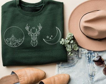 May include: A green sweatshirt with a white graphic design of a mountain, a deer, and a crescent moon. The sweatshirt is folded and laid on a light brown surface. There are also a pair of brown leather mules, a pair of blue denim shorts, a brown fedora hat, and a pair of brown sunglasses.