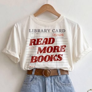 May include: A cream-colored short-sleeve t-shirt featuring a graphic of a library card with the text "READ MORE BOOKS" in red, paired with light blue denim shorts and a brown leather belt with a round buckle, all displayed on a hanger.