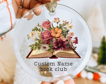 May include: A clear, round glass ornament with a floral design featuring an open book and a bouquet of roses, orange, and burgundy flowers. The text "Custom Name book club" is printed below the book. A gold ribbon is attached for hanging.