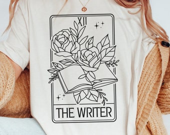 Puede incluir: Camiseta de color marfil con un diseño de carta de tarot en blanco y negro. La carta representa un libro abierto con rosas y el texto "THE WRITER". La parte superior tiene cuello redondo y mangas cortas. La camiseta es de material suave.
