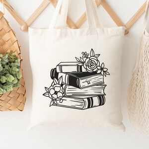 Könnte beinhalten: Cremefarbene Tragetasche mit einer schwarz-weißen Illustration eines Bücherstapels und Blumen. Die Tasche hängt an einer hölzernen Wandgarderobe, neben einem geflochtenen Korb mit Grün und einer Netztasche.