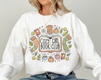 May include: White sweatshirt with a colorful graphic that reads "COZY GIRL BOOK CLUB." The design includes a book, scarf, pumpkins, coffee cup, and leaves. The sweatshirt is a long-sleeved crew neck.