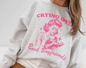 Pode incluir: Um moletom cinza com o texto rosa "Crying Over Book Boyfriends" e uma ilustração rosa de uma mulher lendo e chorando. O moletom é de estilo casual.