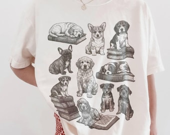 May include: Cream-colored t-shirt featuring a graphic of various dog breeds, each depicted in a detailed, vintage-style illustration. The dogs are positioned on and around stacks of books, creating a charming, literary theme.