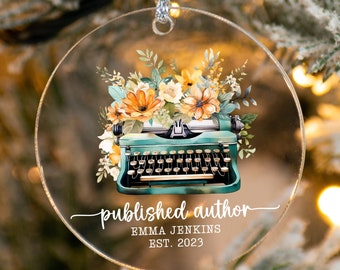May include: A round, clear acrylic ornament with a vintage typewriter and floral design. The text "published author" is written in cursive script, followed by "Emma Jenkins EST. 2023".
