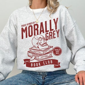 Puede incluir: Sudadera gris con un dise&ntilde;o gr&aacute;fico rojo que dice "Proud Member of the Morally Grey Book Club". El gr&aacute;fico tambi&eacute;n incluye una calavera y huesos cruzados y el texto "Enemies to Lovers Month".