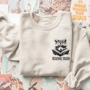 May include: Sand-colored sweatshirt featuring a black and white illustration of a raccoon reading a book. Below the raccoon, the text reads "READING TRASH". The sweatshirt has a crew neck and long sleeves.